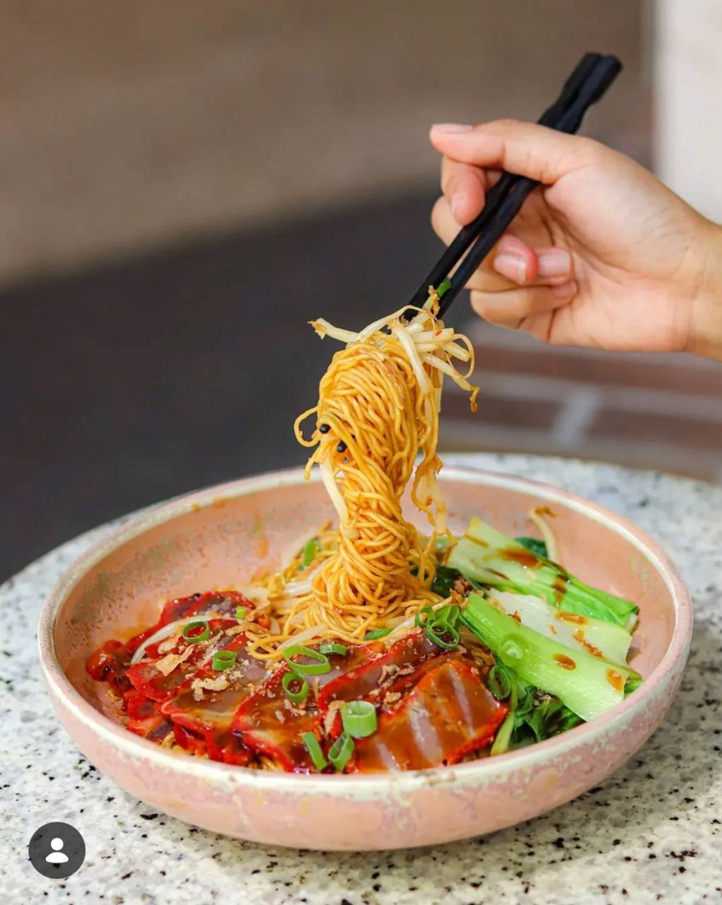 Chopsticks lifting noodles above sliced roast pork and greens at LÜCHA